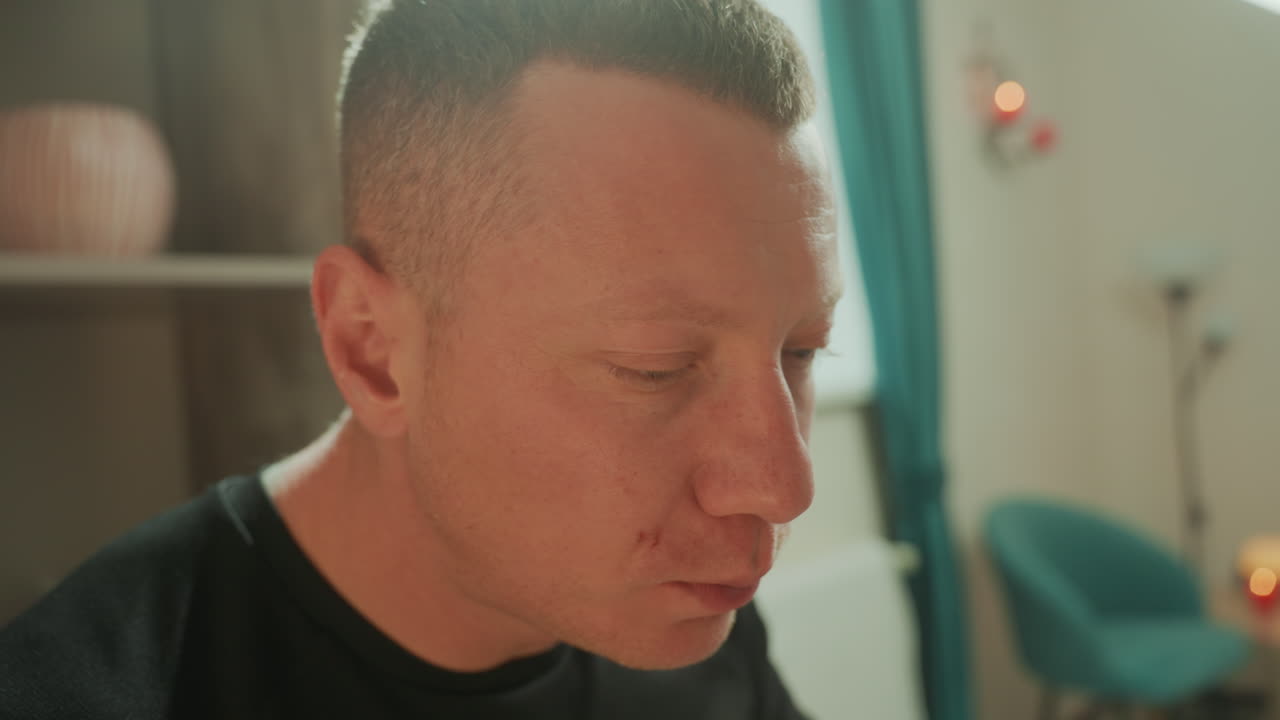 Uomo bianco che riceve un massaggio facciale a microcorrente in una stanza terapeutica illuminata dal sole, occhi chiusi, espressione calma, poltrona color verde acqua, mensola decorativa, candele accese, scatto ravvicinato di profilo che cattura la texture della pelle.