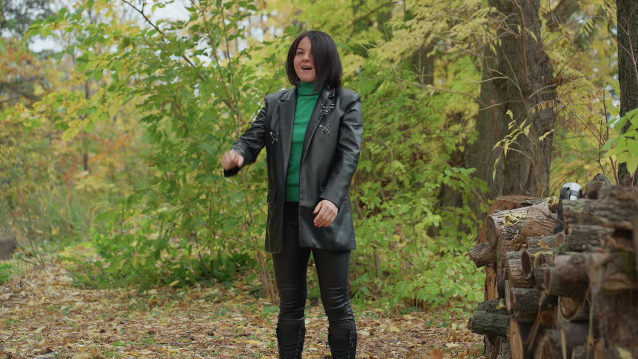 white woman in forest wearing leather green sweater black jacket leather pants standing beside stacked logs and thermos, autumn leaves underfoot, expressive arm raise, singing gesture, confident