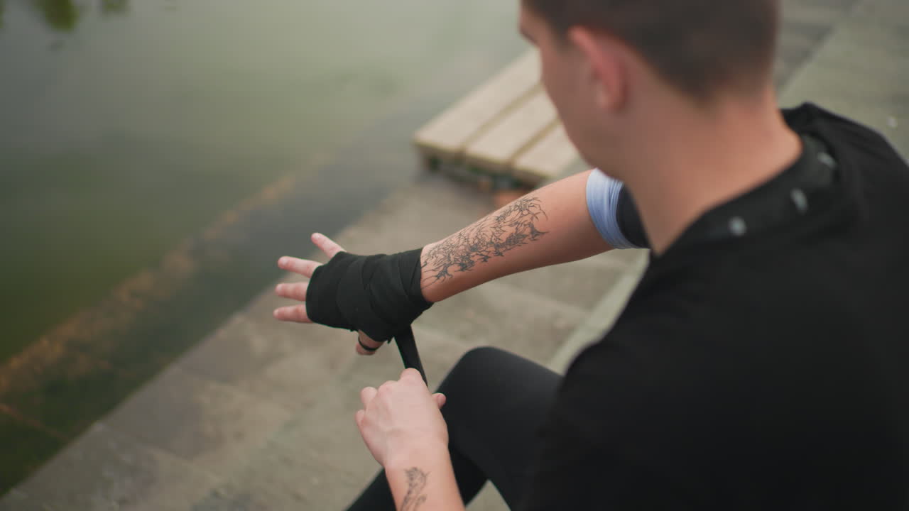 Uomo bianco che fascia le mani vicino a uno stagno, avambracci tatuati, fasce nere, seduto su gradini di cemento, un pugile che si riscalda alla luce della sera, un combattente di strada che si prepara, allenatore che fascia, sparring amatoriale, solitario