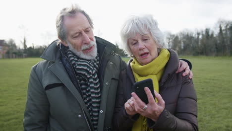 Senior-Couple-Looking-At-Mobile-Phone-On-Autumn-Or-Winter-Walk-Through-Park-Together
