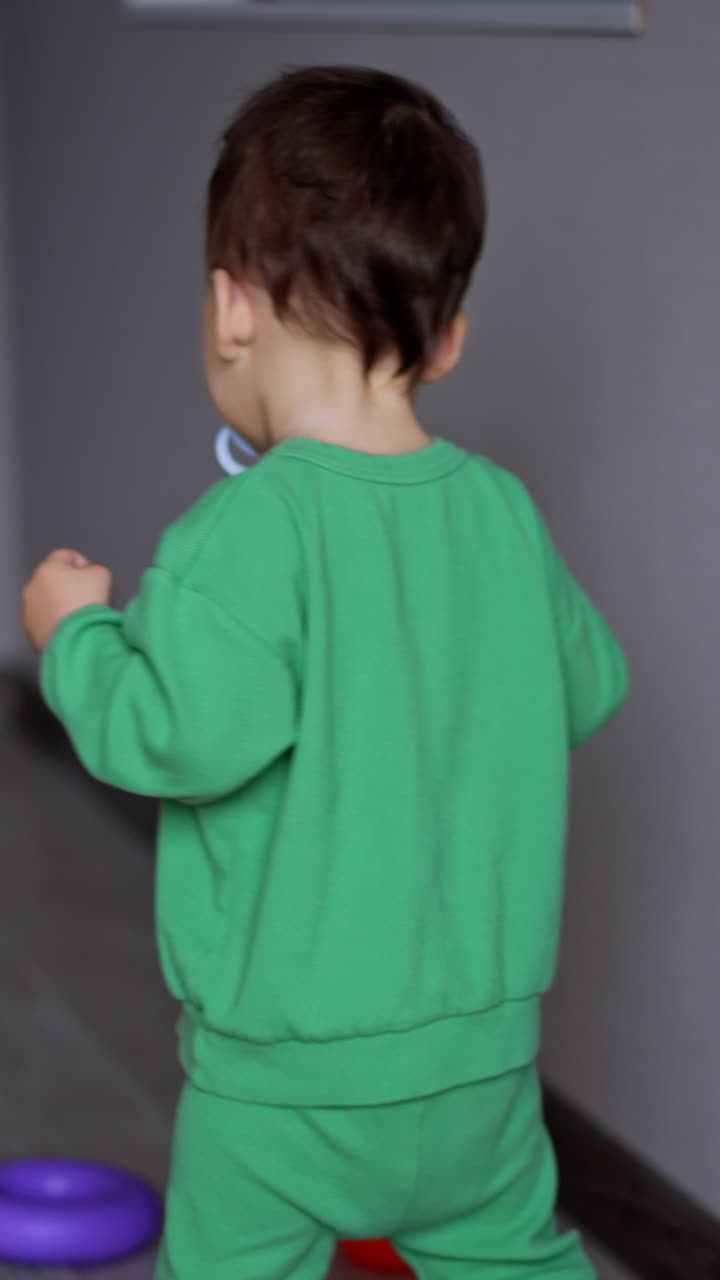 Rear view of a little child in light green sport suit walking by the room. Kid sits to pick up the toys from the floor. Vertical video