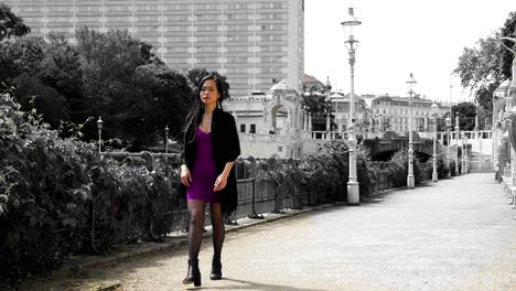 young asian woman in purple dress, black coat walking to the left side of the frame along the wien river at stadtpark, a historical landmark featuring statues of famous viennese composers in austria