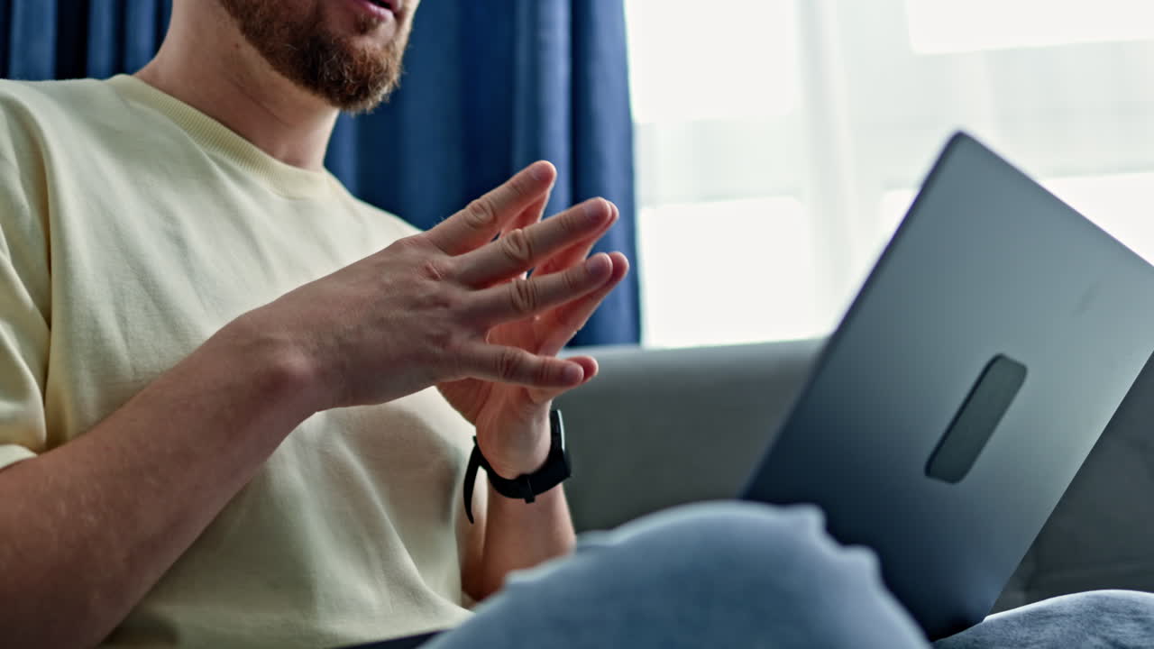 Uomo seduto su un divano che lavora al laptop mentre parla e gesticola con le mani in una stanza con luce naturale che entra da una finestra