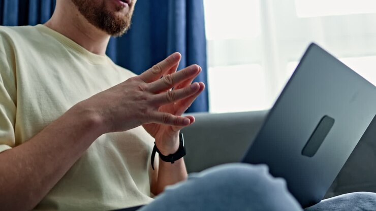 Uomo seduto su un divano che lavora al laptop mentre parla e gesticola con le mani in una stanza con luce naturale che entra da una finestra