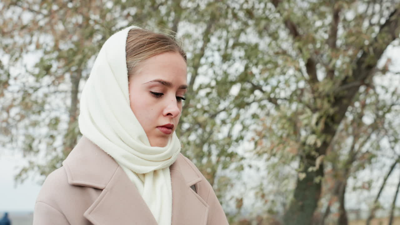 Donna in cappotto sotto un cielo nuvoloso. Donna contemplativa in cappotto beige all'aperto tra betulle. Giovane donna pensierosa con sciarpa che cammina lentamente sotto un cielo nuvoloso circondata da betulle.