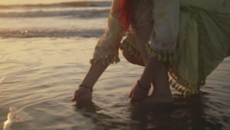 woman squatting in water and feeling it with her hand as the small waves break against the shore