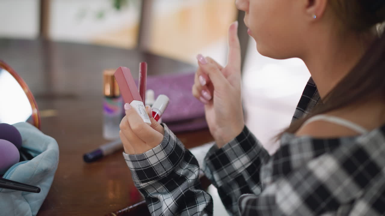 vista parcial de una mujer sosteniendo múltiples artículos de maquillaje mientras señala hacia ellos, contemplando cuál usar fondo incluye productos de belleza en una mesa de madera, bolsa de maquilaje y espejo
