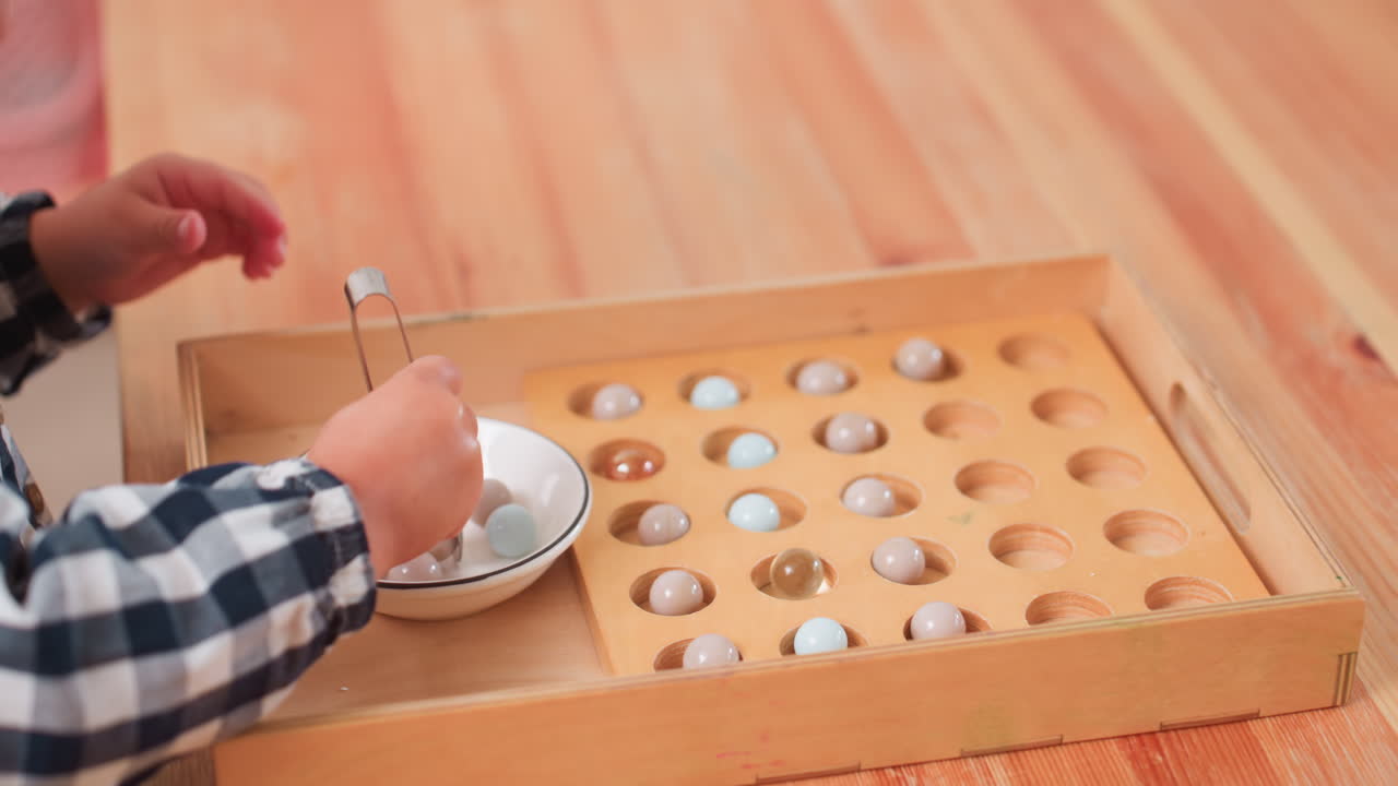 Close up of little child with blonde hair in checkered shirt concentrating while placing colorful balls into wooden puzzle box on table, focused on fine motor skills during indoor learning activity