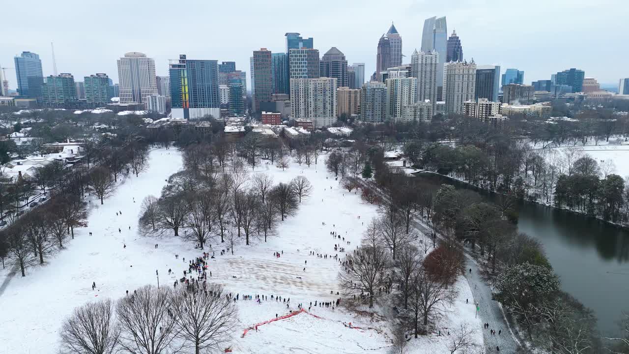 Static aerial wide shot of people sledding in a snow covered Piedmont Park with Midtown Atlanta buildings in the background.