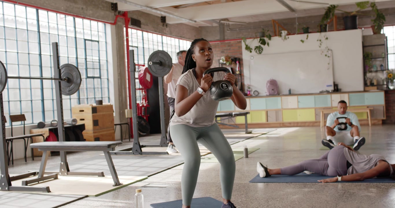 Corso di ginnastica diversificato, donna afroamericana che esegue uno squat goblet su indicazione, sollevando un kettlebell per la tonificazione.
