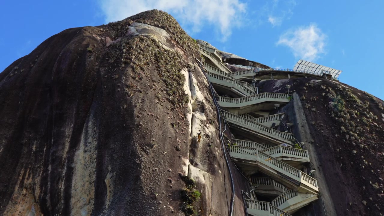 4K low angle static shot of the famous zig-zagging stairs built into the granite cliff of El Penol rock under a blue sky.