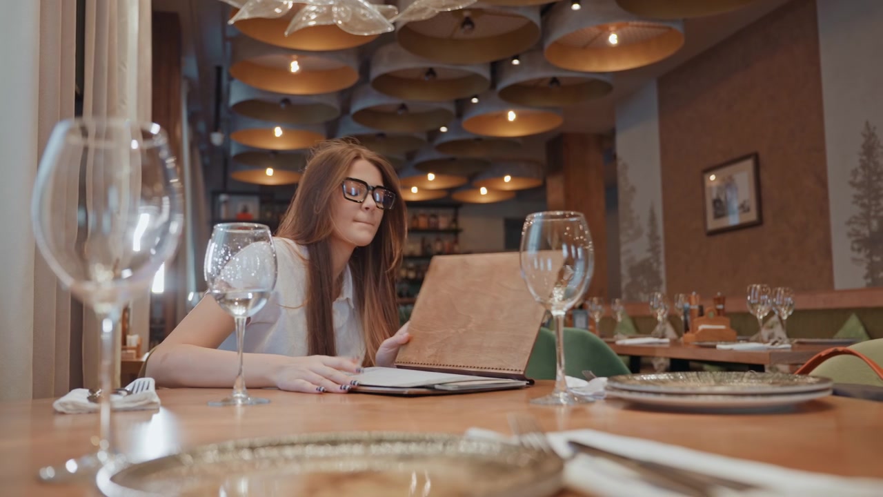 Ung kvinde læser menu på restaurant, glas og lysekrone i hyggeligt interiør, bladrer sider, nipper til et glas, tjekker telefon, venter på selskab, bliver træt, hovedet hviler på bordet.