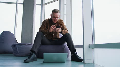 young man with gadgets on windows background. handsome guy is sitting on ottoman in office and using mobile phone. laptop on the floor. freelance concept.