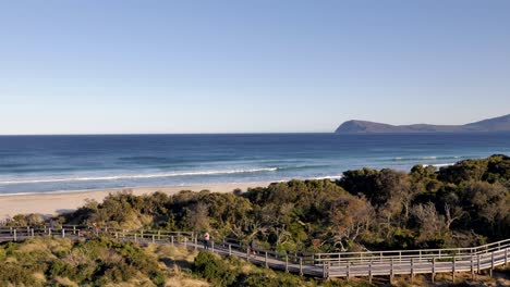 beach waves roll in as tourists walk along boardwalk among bush landscape coastline during evening sunset on bruny island neck isthmus tasmania, australia