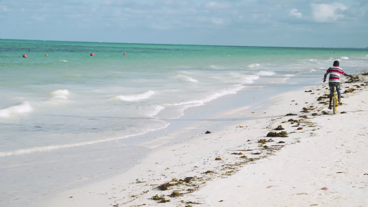 Black kid pedaling on monocycle by sea waves on sand beach in Zanzibar