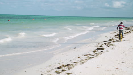 Black-kid-pedaling-on-monocycle-by-sea-waves-on-sand-beach-in-Zanzibar