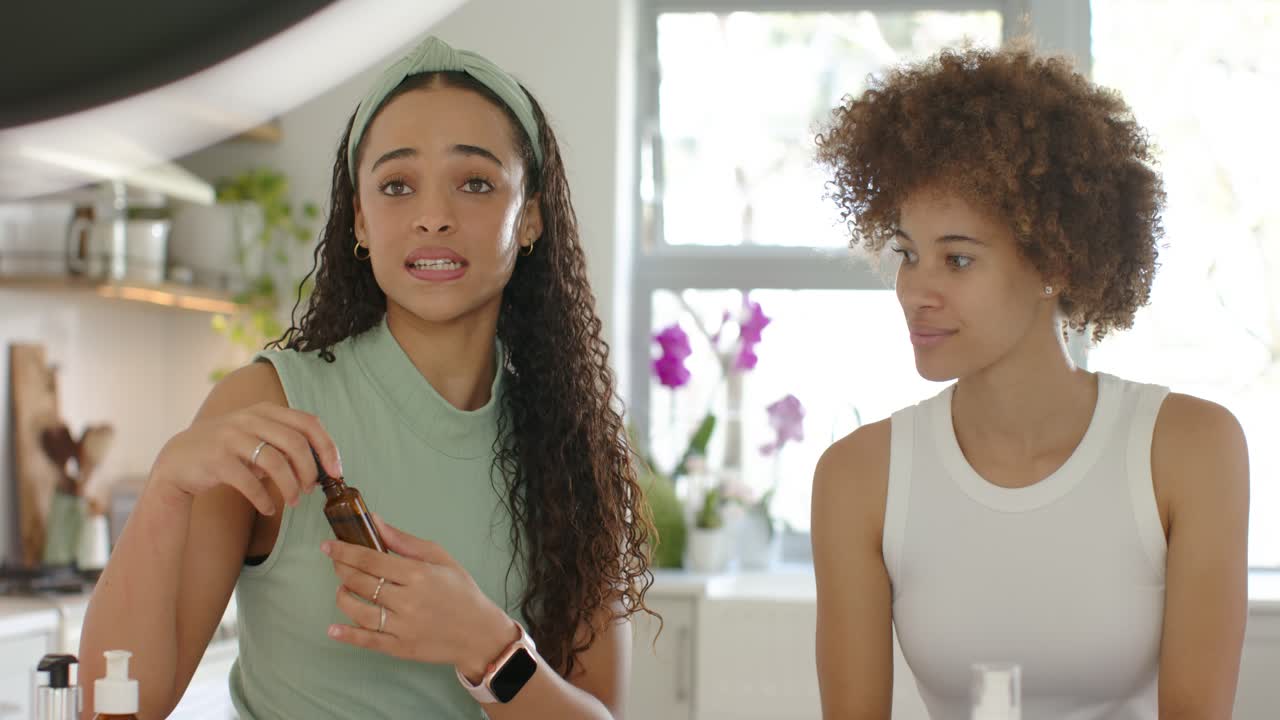 African American women left woman opening amber dropper showing in kitchen while right trying skin
