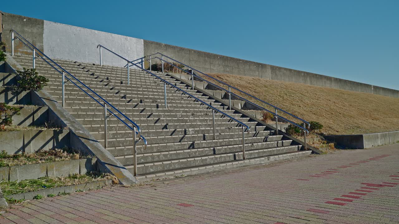 Una toma amplia de escaleras de hormigón con pasamanos plateados que suben por un terraplén cubierto de hierba. Las sombras caen sobre los escalones bajo un cielo azul claro.