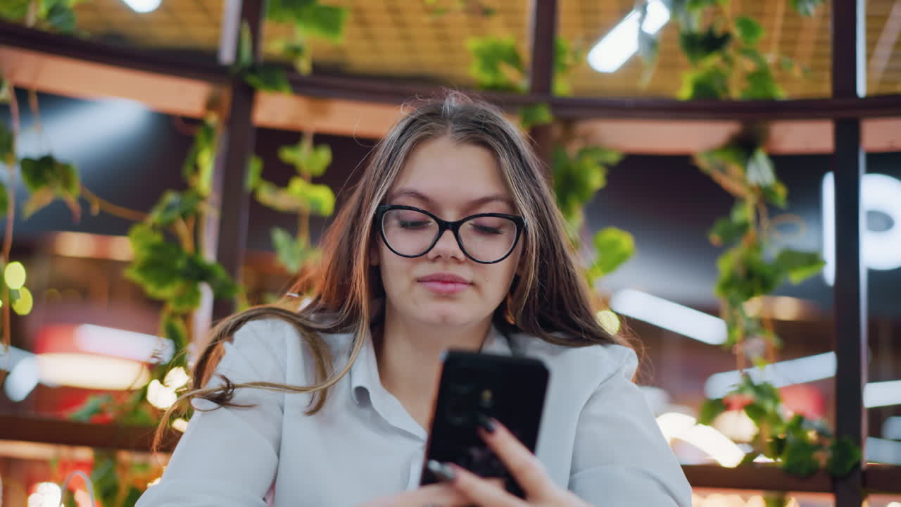 primer plano de una dama elegante con gafas, centrada en operar su teléfono, con luces bokeh suaves que mejoran el fondo cálido, mostrando una mezcla de uso de tecnología y ambiente elegante