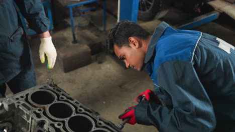 automotive engineer in blue uniform loosening a bent nut with red gloves while observer in white gloves shows him something in an automotive workshop