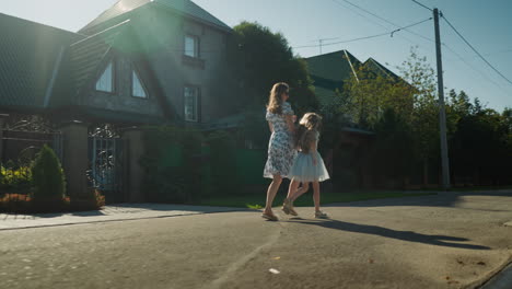 back view of woman in floral dress running hand in hand with young girl as sunlight casts soft silhouette effect on quiet residential street near large house with green rooftops and ornamental gate