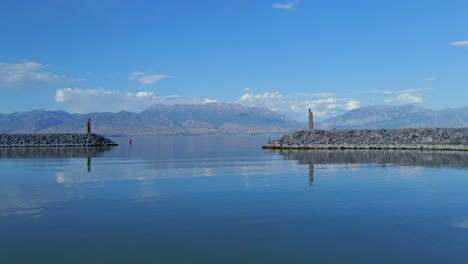 Toma-Aérea-De-La-Entrada-Al-Puerto-Deportivo-De-Saratoga-Springs-En-El-Lago-Utah-Desde-El-Final-De-Un-Pontón-Rojo-Siguiendo-Las-Boyas-Del-Canal-Con-Montañas-Distantes-En-El-Fondo-Reflejándose-En-Las-Aguas-Tranquilas