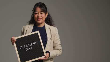 studio portrait of female teacher standing against grey background holding notice board reading teachers day