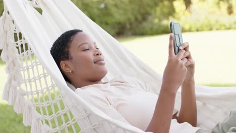 happy african american woman lying in hammock on terrace, using smartphone, slow motion