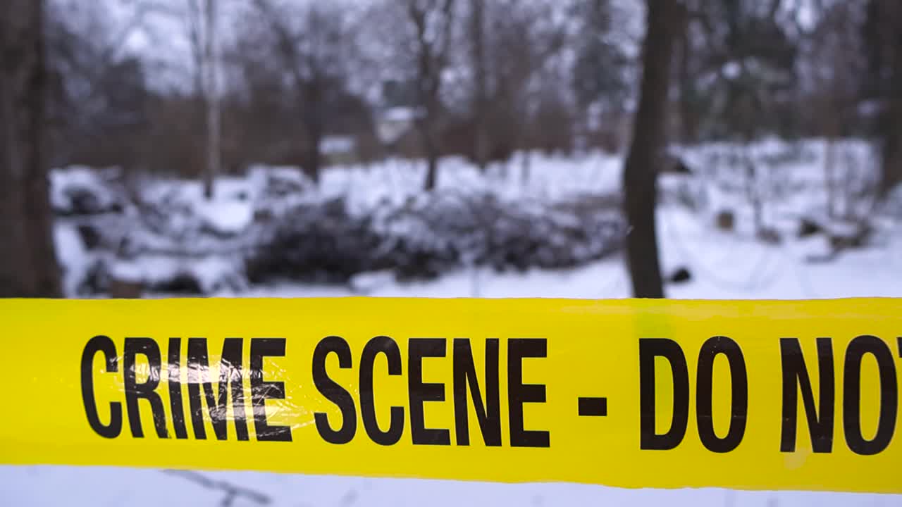 Close up glide along yellow crime scene tape with shallow depth of field, set against snowy woodland and bare winter trees during a darkening, moody investigation atmosphere and softly falling snow