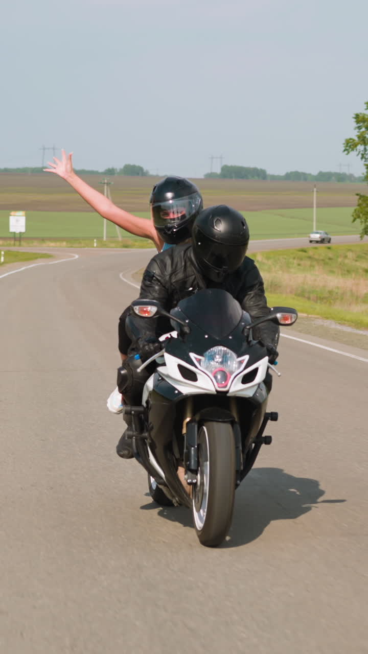 Joyful woman with raised arm enjoys motorcycle riding with boyfriend along road past green trees and empty field at countryside slow motion