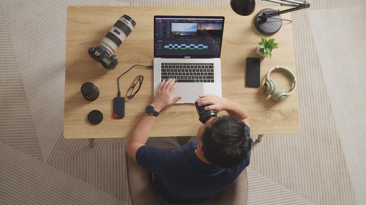 vista superior de un editor masculino asiático bebiendo café mientras está sentado en el espacio de trabajo usando una computadora portátil junto a la cámara editando el video en casa