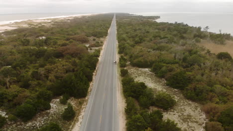 Aerial-pull-back-from-two-bikers-enjoying-a-ride-down-a-very-long-and-straight-road,-to-a-panoramic-view-of-the-woods-and-dunes-of-the-Jersey-Shore