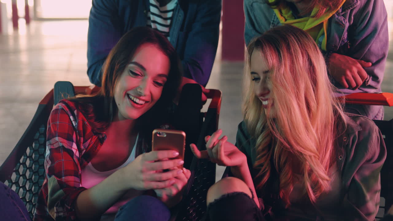vista de cerca de dos niñas caucásicas sentadas en un carrito y una niña mostrándole a la otra algo en la pantalla de su teléfono inteligente mientras se ríen