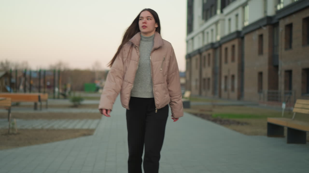 young girl in a peach jacket and black trousers is rollerblading along a paved path in a serene urban park. The scene captures the peaceful atmosphere with benches and people sitting in the background