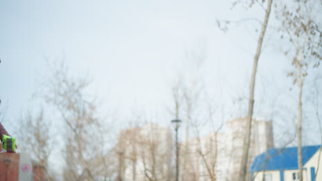 black guy wearing dark gray hoodie holding orange gloves facing woman in pink jacket and beanie walking outdoors in park during early spring, bare trees and urban buildings in background