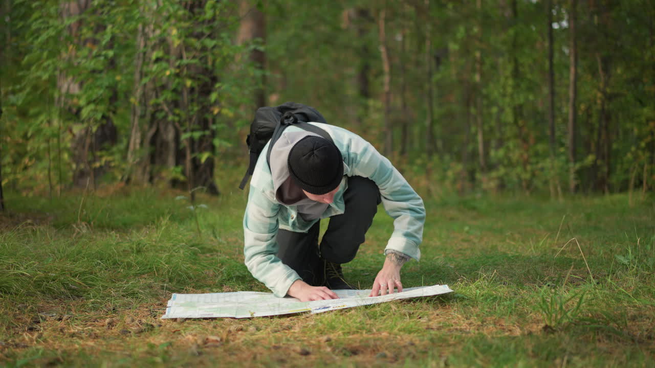 male hiker kneeling studying map, backpacked traveler examining routes on ground, forest clearing setting with trees and grass, focused route plotting and waypoint checking, sense of determination