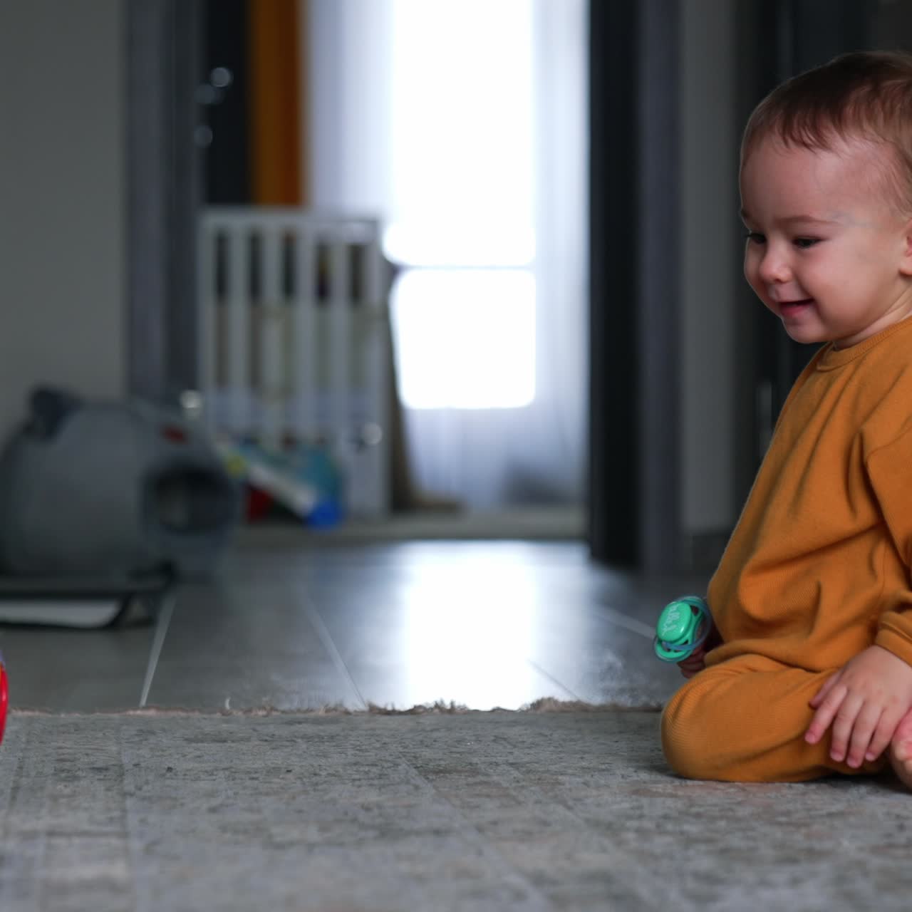 Sweet kid sitting on the floor pushes a ball watching it roll. Nice little kid playing with toys in the room. Blurred backdrop