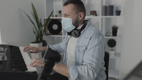 male musician with medical face mask playing electric keyboard and using a laptop at home during lockdown due to the covid 19 pandemic