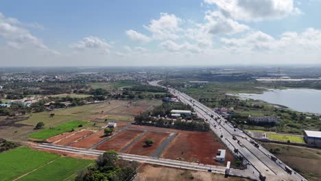 sweeping drone shot captures a bustling highway traversing varied landscapes—lush fields, emerging construction, and tranquil water bodies under a partly cloudy sky. new plots for sale