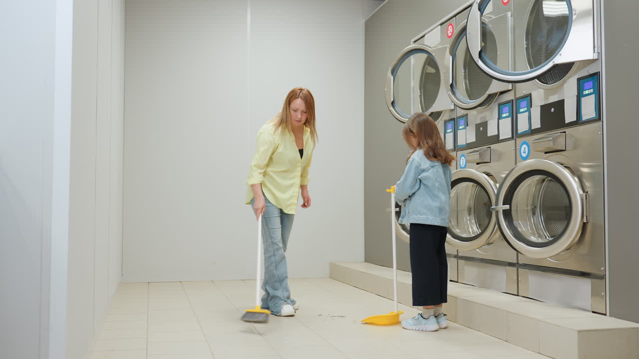 Toddler holds dustpan while mother sweeps tiled floor inside bright laundromat near industrial washers, family chore scene showing hygiene, responsibility, guidance, teamwork during routine cleaning