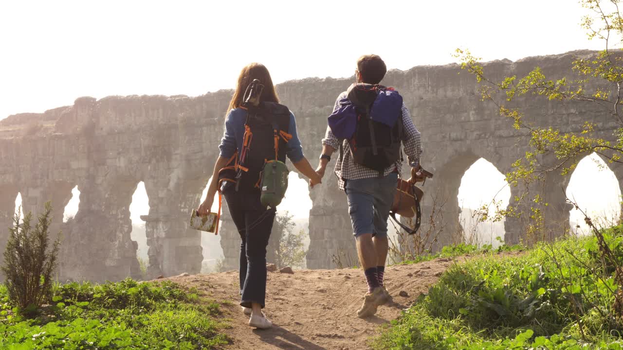 joven pareja encantadora mochileros turistas caminando de la mano hacia el acueducto romano arcos en el parco degli aquedotti ruinas del parque en roma en romántico amanecer brumoso con guitarra y saco de dormir cámara lenta