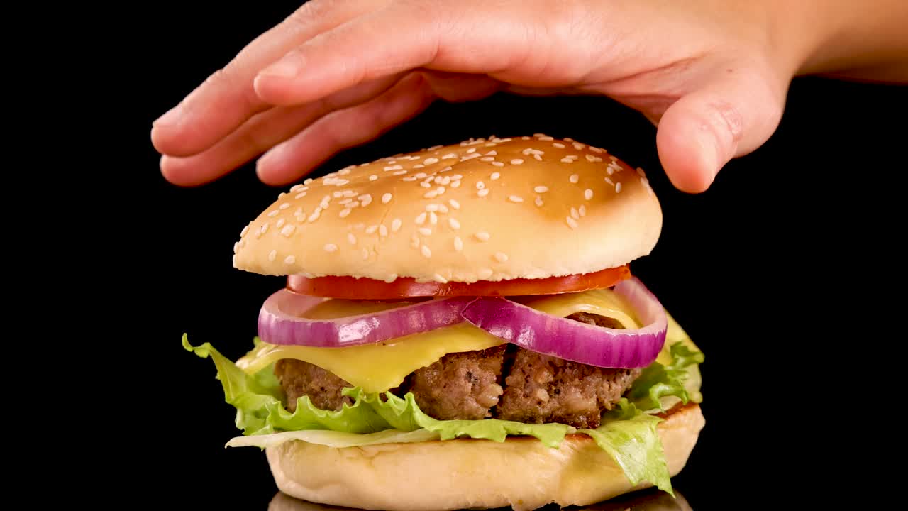 A hand gently places a sesame seed bun atop a beef burger with lettuce, tomato, and onion, isolated on a black background with even lighting