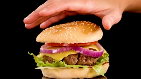 a hand gently places a sesame seed bun atop a beef burger with lettuce, tomato, and onion, isolated on a black background with even lighting