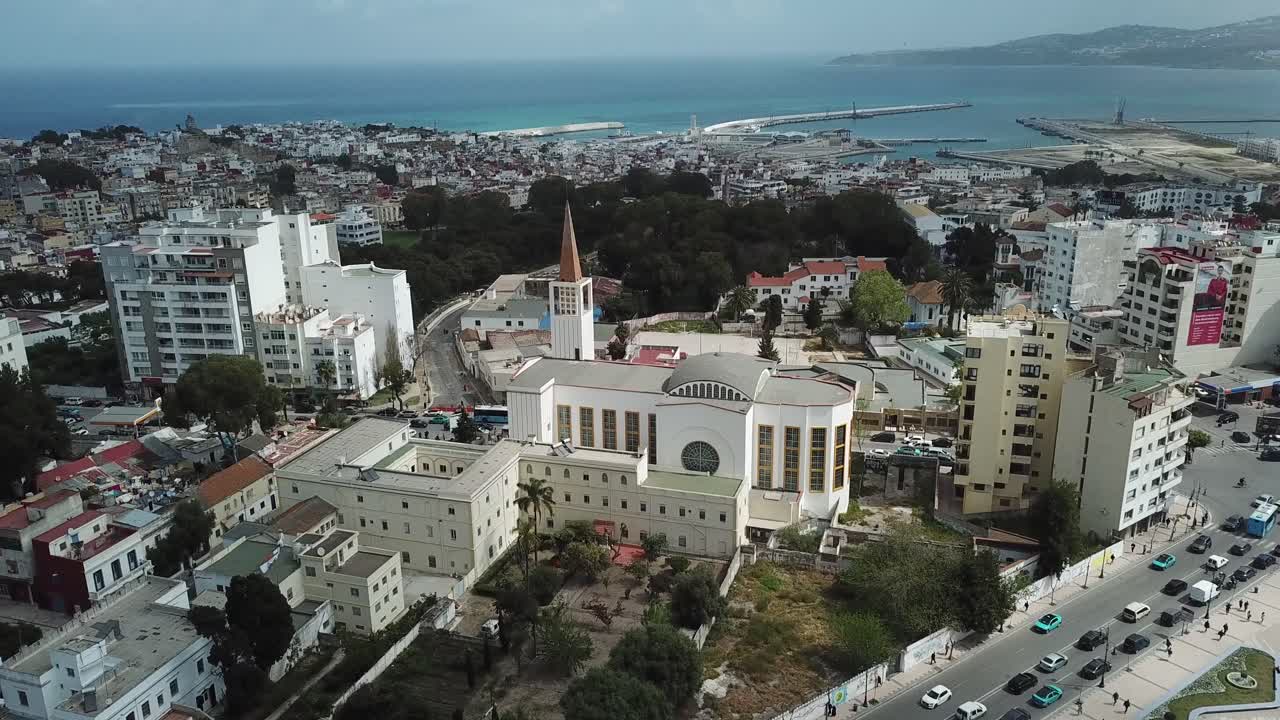Vista aérea cinematográfica de Tánger, Marruecos, mostrando una iglesia histórica, calles urbanas y el puerto mediterráneo bajo un cielo brillante.