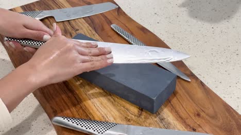 hands methodically sharpen a large chef’s knife on a rectangular whetstone atop a wooden board, under bright, even kitchen lighting with steady overhead camera angle