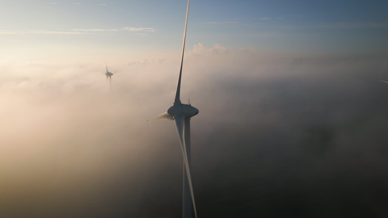 Nebbia e nuvole si muovono tra le pale rotanti di grandi turbine eoliche all'alba.