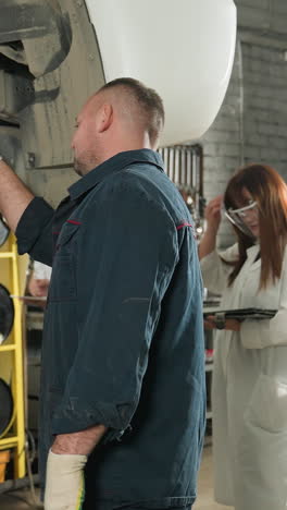 vista trasera de un hombre en uniforme azul con científicos de laboratorio tomando notas en una tableta en un taller mecánico. entorno industrial con herramientas, colaboración, investigación y análisis técnico en progreso
