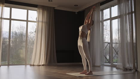 una joven hermosa en ropa deportiva blanca hace estiramiento y yoga en un gimnasio con grandes ventanas en cámara lenta