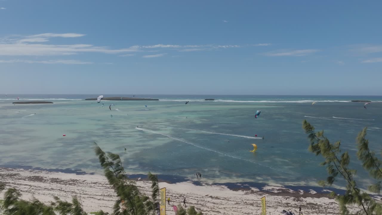 Drone ascends above Sakalava Bay in Madagascar capturing kitesurfers gliding over shallow turquoise waters near the white beach, framed by sparse coastal vegetation under a bright blue sky