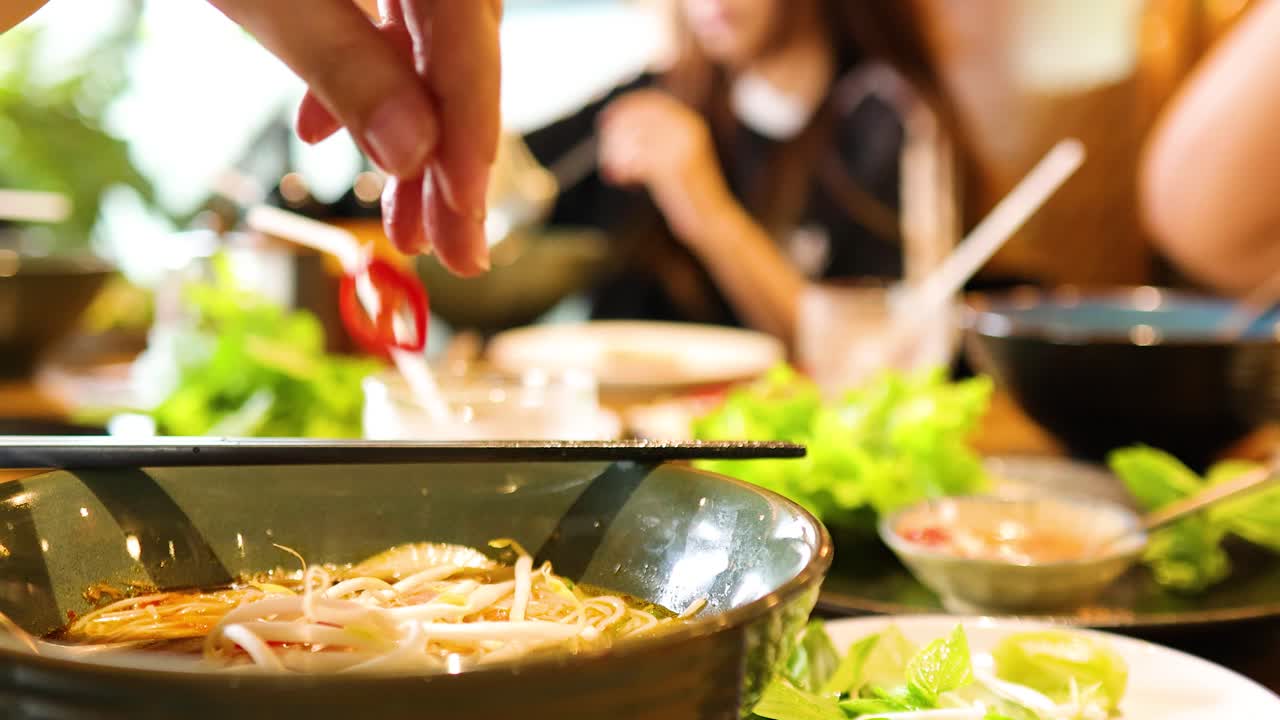 Hands adding ingredients to pho in a lively restaurant setting with warm lighting and vibrant colors